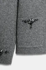 Chaqueta de punto gris con cuello solapa, manga larga y cierre frontal con botones forrados. Presenta bolsillos delanteros de plastrón y detalles de abalorios joya en los bolsillos y en los puños.