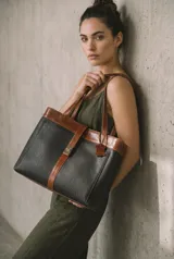 Cartera tipo tote de cuero vacuno, con diseño bicolor en negro y marrón. Presenta doble repartición, cierre frontal con broche metálico y correas de hombro. Incluye etiqueta colgante de cuero y logo grabado en la parte inferior.