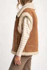 Chaleco estilo gamulán de corte oversize, confeccionado en material tipo gamuza color camel con interior y bordes de corderito sintético color crema. Presenta cierre frontal completo, bolsillos laterales y detalles de tiras decorativas en los hombros.