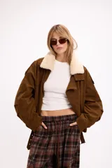 Campera de gamuza color marrón con cuello de corderito sintético en color crema. Presenta un diseño oversize con cierre frontal, solapas con broches metálicos y una tira ajustable en el ruedo.