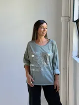 Remera de manga larga color verde, con cuello redondo y diseño holgado. Presenta un detalle frontal con parches de lentejuelas plateadas y estampado tipográfico.