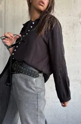 Camisa de color negro con cuello clásico y cierre frontal de botones blancos. Presenta mangas largas con detalles bordados en los puños.