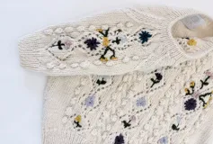 Sweater de manga corta de lana merino color marfil, con cuello redondo, diseño texturizado en relieve y bordados florales multicolores hechos a mano.