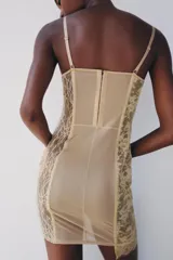Vestido corto semitransparente tipo corsetero color beige, confeccionado con encaje y detalles de varillas interiores que estilizan la figura. Presenta escote redondo, tirantes finos ajustables y bajo asimétrico con una lazada lateral de satén.