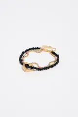 Pulsera dorada de múltiples tiras, combinando una cadena con cristales brillantes, cuentas de vidrio facetadas negras y un dije en forma de corazón con detalles esmaltados y letras grabadas.