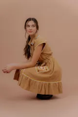 Vestido corto beige de algodón con cuello camisero y cierre frontal de botones. Presenta mangas cortas y detalles bordados a contratono en la zona del pecho y en los bolsillos, además de un volado en el ruedo.