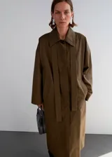 Abrigo largo oversize de algodón color verde militar, con cuello camisero, cierre delantero de botones ocultos, bolsillos laterales y tiras decorativas en el frente.