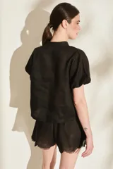 Blusa negra de lino con cuello mao, mangas cortas abullonadas y cierre frontal parcial con botones dorados. Presenta pequeños tajos laterales en el ruedo.