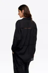 Camisa de corte recto en color negro, con cuello camisero, cierre frontal de botones y manga larga con puños abotonados. Presenta un bolsillo de plastrón en el pecho y detalles de bordados a tono con aplicaciones de encaje tipo entredós distribuidos por toda la prenda.