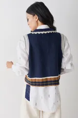 Chaleco de lana color marrón con cuello estilo bebé, cierre frontal con botones grandes y detalle de puntilla en cuello, sisas y bolsillo frontal. Presenta una franja decorativa en color rojo en el ruedo.