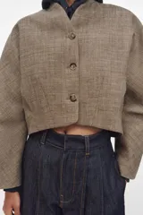 Chaqueta corta de corte estructurado confeccionada en tejido con mezcla de lana. Presenta cuello en pico, cierre frontal con botones y mangas largas. El diseño incluye bolsillos delanteros y un forro interior a contraste.