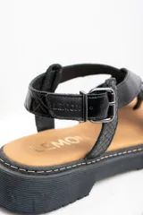 Sandalia de cuero color negro charol, con tiras cruzadas y hebilla ajustable al tobillo. Plataforma con suela track.
