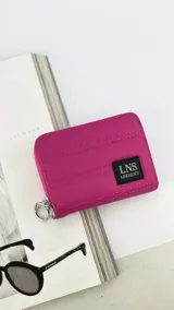 Billetera corta de diseño capitoneado en color fucsia, con cierre perimetral y tirador metálico circular. Presenta un parche rectangular con el logo de la marca LNS Lincoln's en el frente.