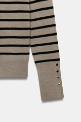 Jersey de punto liso en color beige, con cuello alto y manga larga con detalle de botones metálicos en los puños.