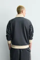 Sudadera de corte relajado en color azul marino con cuello alto y cierre frontal de cremallera. Presenta bandas laterales en color crema a contraste que recorren las mangas, puños y bajo en tejido rib, y bolsillos laterales de vivo.