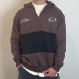 Canguro con capucha y cierre frontal parcial, de diseño color block en tonos marrón y negro. Presenta estampado gráfico en el pecho con el logo de la marca y detalles de diseño.