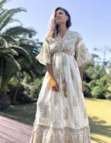 Túnica larga de estilo boho, color crudo con estampado de arabescos y detalles en hilo dorado. Presenta escote en V, mangas amplias tipo kimono y cordón ajustable en la cintura con borlas decorativas.