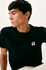 Remera negra de manga corta con cuello redondo, confeccionada en algodón. Presenta un pequeño parche rectangular con el logo de la marca en el pecho.