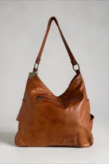 Cartera de cuero natural en color marrón, con diseño envolvente, caída suave y bolsillo exterior cruzado con el nombre de la marca bordado. Cuenta con tiras de hombro, herrajes metálicos y un dije colgante geométrico.