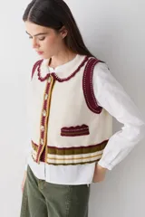 Chaleco de lana color marrón con cuello estilo bebé, cierre frontal con botones grandes y detalle de puntilla en cuello, sisas y bolsillo frontal. Presenta una franja decorativa en color rojo en el ruedo.