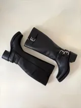Bota de caña alta estilo biker, confeccionada en cuero negro. Presenta tres hebillas metálicas decorativas, cierre lateral y taco de madera.