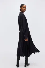 Vestido camisero midi negro de corte holgado, con cuello, escote en V y cierre frontal de botones dorados. Cuenta con mangas largas abullonadas con puños anchos y un lazo ajustable para atar en la cintura.