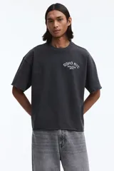 Remera boxy de algodón gris oscuro, con corte oversized, hombros caídos, cuello redondo ribeteado y estampa pequeña en el pecho con texto en blanco.