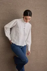 Blusa de lino color blanco con cuello mao, cierre frontal con botones, mangas largas abullonadas con puños ajustados y detalle de volado en el ruedo.