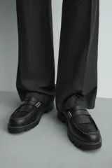 Mocasín negro de cuero liso con antifaz y flecos en el empeine. Presenta costuras blancas visibles y suela track gruesa de goma.