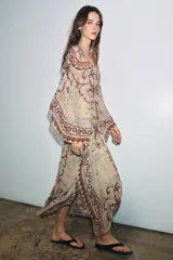 Vestido tipo túnica de viscosa con escote en pico y mangas tipo capa. Presenta un diseño estampado con motivos ornamentales en tonos tierra, beige y rosado, con pliegues en la cintura y bajo asimétrico.