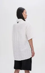 Camisa de manga corta en color off-white, con cuello clásico y cierre frontal de botones. Presenta un diseño con textura de rayas verticales y motivos circulares.