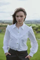 Camisa blanca de corte clásico con cuello camisero, mangas largas abullonadas y detalle de volados en el canesú delantero. Presenta cierre frontal con botones.