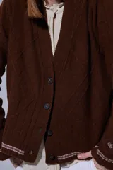 Saco tejido en lambswool con diseño de rombos y trenzas en el frente. Presenta cierre frontal con botones, cuello en V y detalles de rayas en contratono en puños y ruedo. Incluye pin bordado en la manga.