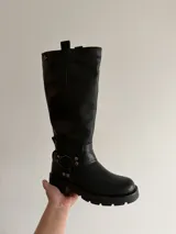 Bota de caña alta estilo biker, confeccionada en cuero sintético negro. Presenta una suela track con plataforma, puntera redondeada y un detalle de arnés con hebilla metálica y tachas en la zona del tobillo.