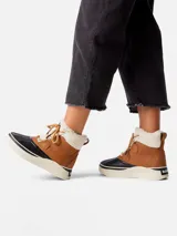 Bota de estilo pato Sorel, confeccionada en cuero impermeable color tostado con forro interior de borrego sintético. Presenta una carcasa de goma vulcanizada negra en la base, entresuela de plataforma en color crema y cierre con cordones.