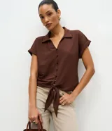 Blusa color marrón con cuello solapa, mangas cortas con vuelta y cierre frontal de botones. Cuenta con un diseño texturizado y un lazo para anudar en el bajo.