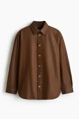 Camisa de corte oversized en denim de algodón rígido revestido, color marrón oscuro. Presenta cuello inglés, tapeta clásica con botones de metal y canesú en la espalda. Tiene hombros caídos, mangas largas con puños abotonados y bajo redondeado.