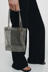 Bolso tipo tote cuadrado, confeccionado en tejido de malla metálica semitransparente. Cuenta con dos asas superiores fijas y detalles de remaches metálicos en la unión de las asas con el cuerpo del bolso.