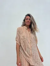 Camisa de corte oversize y manga corta, confeccionada en tela ligera color beige con rayas verticales bordadas en color crudo. Presenta cuello mao, cierre frontal con botones y bolsillos frontales.
