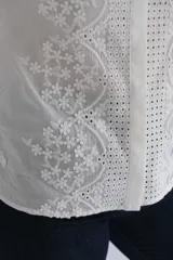 Blusa blanca de manga larga con cuello mao y cierre frontal de botones ocultos. Presenta un delicado diseño con bordados florales y detalles calados verticales en el frente.