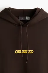 Canguro marrón de peso medio, con capucha de doble capa con cordón de ajuste, bolsillo canguro frontal y estampa con la palabra "OBSESSED" en amarillo en el pecho. Presenta puños y bajo acanalados.