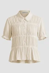 Camisa Belen de manga corta abullonada, color beige, confeccionada en tejido liviano con textura. Presenta cuello redondo con solapa y delicadas franjas horizontales de puntilla y encaje, con cierre frontal de botones.