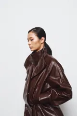 Chaqueta amplia de efecto piel en color marrón oscuro, con cuello alto, manga larga y bolsillos delanteros. Presenta cintura ajustable, forro interior y cierre frontal con cremallera y botones ocultos por solapa.
