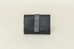 Billetera pequeña de cuero sintético negro con cierre de solapa y broche a presión. Presenta un aplique de cuero gris en la solapa y costuras a contratono.