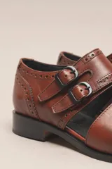 Zapato tipo monk strap con aberturas laterales, confeccionado en cuero vacuno metalizado color bronce. Presenta detalles de perforaciones estilo brogue en la puntera y el talón, y doble hebilla metálica en el empeine.