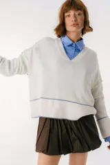 Sweater de punto color blanco con cuello en V, de corte holgado y manga larga. Presenta detalles de costuras y bordes en color azul contrastante en el ruedo y los puños.