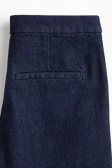 Pantalón de jean azul oscuro, tiro alto, con corte ajustado hasta la rodilla y pierna acampanada (bootcut). Presenta cierre frontal con botón y corchete ocultos, bolsillos al bies y bolsillos decorativos traseros.