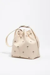 Bolso tipo saco color beige de piel vacuna, con estampado de palmeras pequeñas en marrón, cordón de ajuste frontal y asa regulable con hebilla metálica. Incluye el logo grabado de la marca en la parte inferior.