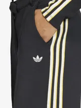 Pantalón de corte holgado y estilo sastrero, confeccionado en tejido de doble punto color negro. Presenta las icónicas tres tiras de la marca en los laterales en combinación de colores blanco y amarillo, con el logo del trébol bordado en la parte frontal. Cuenta con cierre de cremallera y bolsillos laterales.