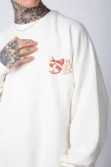 Buzo de tejido friza color crema, de corte oversize y cuello redondo. Presenta un pequeño bordado o parche en el pecho izquierdo con un diseño de cabeza de animal en tonos rojizos/terracota.